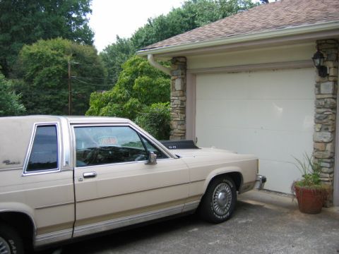 1985 Ford LTD Crown Victoria - A tan sedan with a hood scoop is parked in front of a closed garage door.