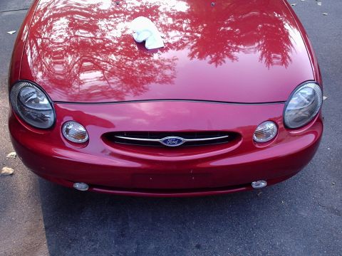 1998 Ford Taurus - Elevated head-on view of the front end of a dark red Ford sedan, showing the small oval Ford blue-oval grille badge, clear oval headlamps w…