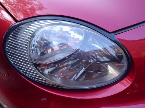 1998 Ford Taurus - Close-up of a clear oval composite headlamp assembly set into dark red bodywork, showing the multi-faceted reflector, integrated ribbed sid…