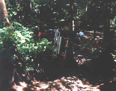 1992 Ford Bronco - An off-road vehicle has rolled onto its side in a wooded area, with two individuals observing the scene.