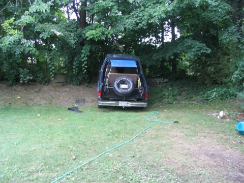 1986 Ford Bronco - Rear view of a 1986 Ford Bronco parked in a yard, showing the spare tire and an open top covered by a blue tarp.