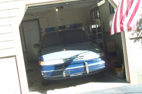 1991 Ford LTD Crown Victoria - A blue police car with a light bar is parked inside a garage, with an American flag visible on the right.