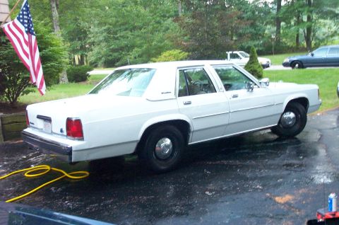 1991 Ford LTD Crown Victoria - A white sedan is parked outside with an American flag. Another vehicle is visible in the background.