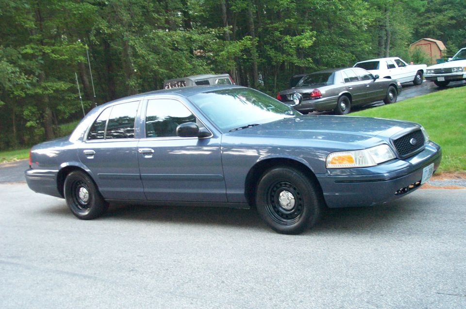 Blue Ford Crown Victoria parked on a street with trees in the background. Other cars are visible on a grassy incline
