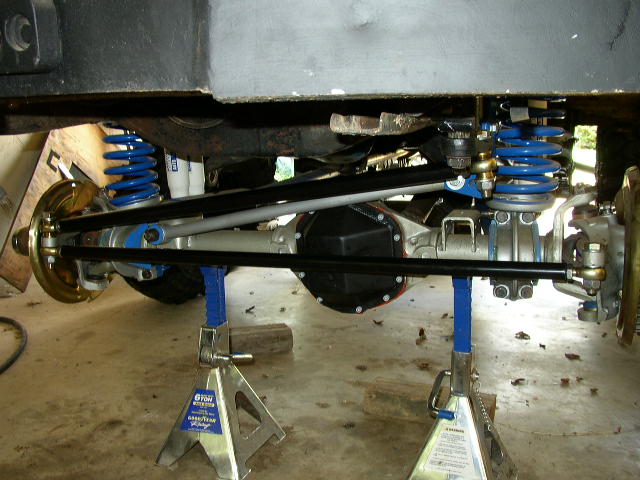 Underneath view of a vehicle's rear axle, differential, and suspension components supported by jack stands. Blue coil springs are visible