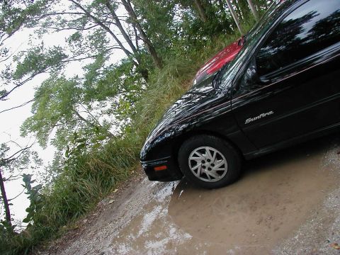 1997 Ford Taurus - A black Pontiac Sunfire is parked on a muddy road next to a red car, surrounded by trees.