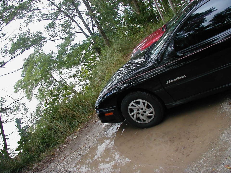 Black Pontiac Sunfire with red car behind it, parked on a muddy road with trees and water