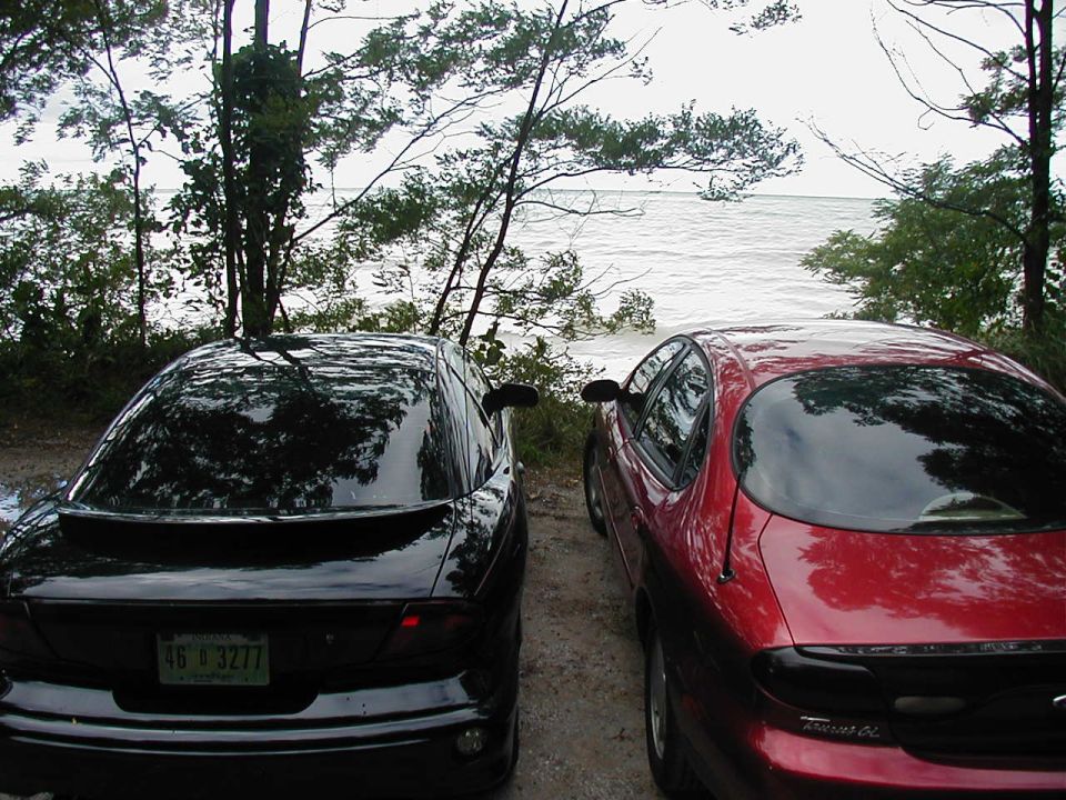 Two sedans, one black and one red, parked on a dirt road next to a large body of water with trees in the background