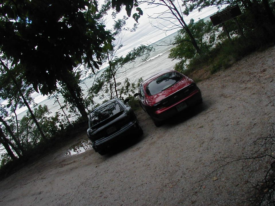 Two sedans, a black one and a red one, parked on a dirt road with a body of water in the background. The red car is a 1997 Ford Taurus