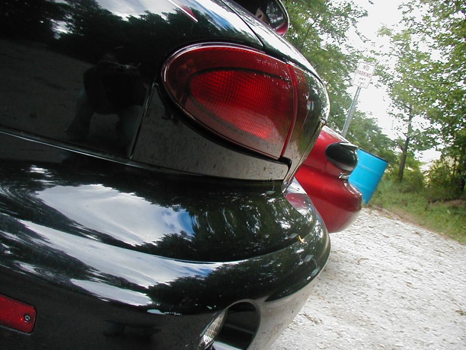 Black Pontiac Sunfire rear end, red car behind it, blue barrel nearby