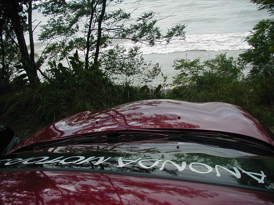 Red car hood and windshield with trees and ocean view. Text reads 'ANONDA MOTORS'