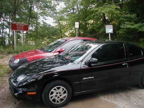 1997 Ford Taurus - Two cars are parked on a dirt road near signs in a wooded area.