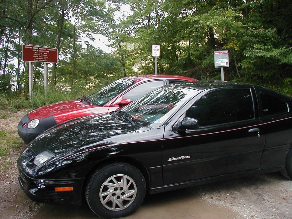 Black Pontiac Sunfire coupe and red car parked on dirt road with trees and signs