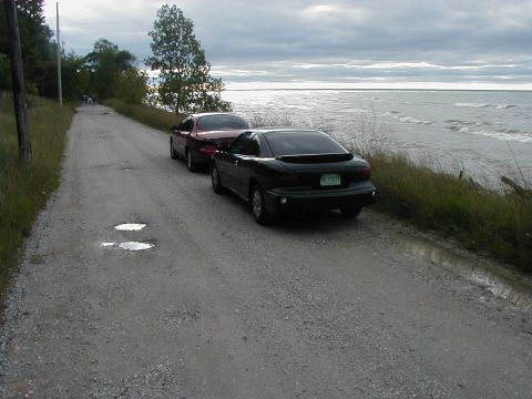 1997 Ford Taurus - Two cars are parked on a gravel road beside a large body of water under a cloudy sky.
