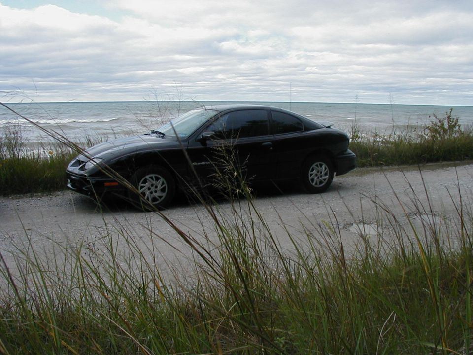 Black coupe parked on a dirt road by the water, with tall grass in the foreground