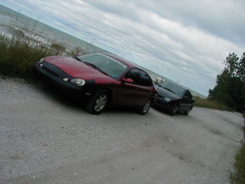 1997 Ford Taurus - A red 1997 Ford Taurus is parked on a dirt road near the water, with another dark car behind it.