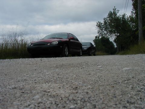 1997 Ford Taurus - Two sedans are parked on a gravel road under a cloudy sky.