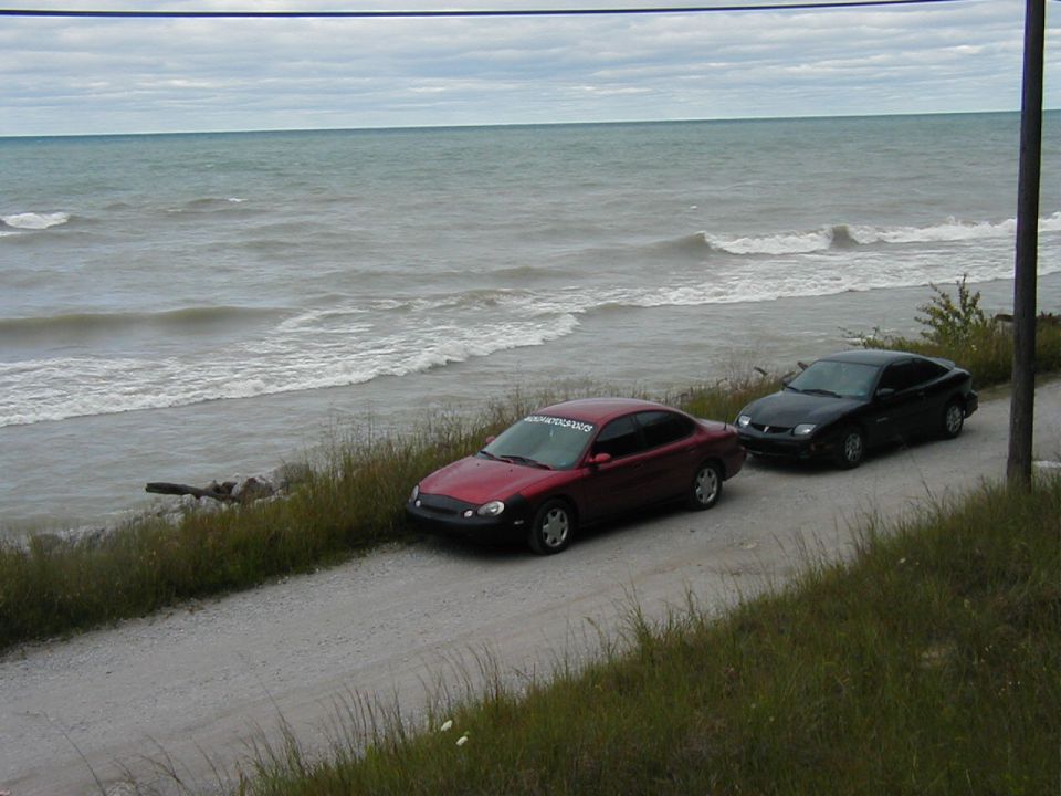 Red sedan and black sedan parked on a gravel road near the ocean