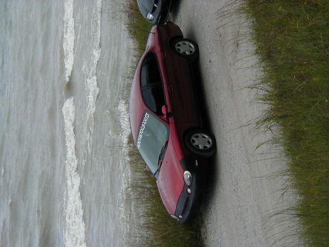 1997 Ford Taurus - A red sedan is parked on a dirt road beside a body of water, with another vehicle partially visible.