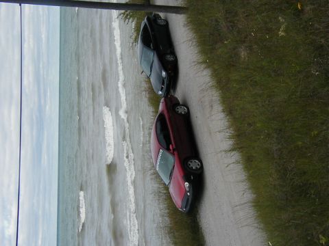 1997 Ford Taurus - Two cars are parked on a dirt road overlooking the ocean with waves crashing on the shore.