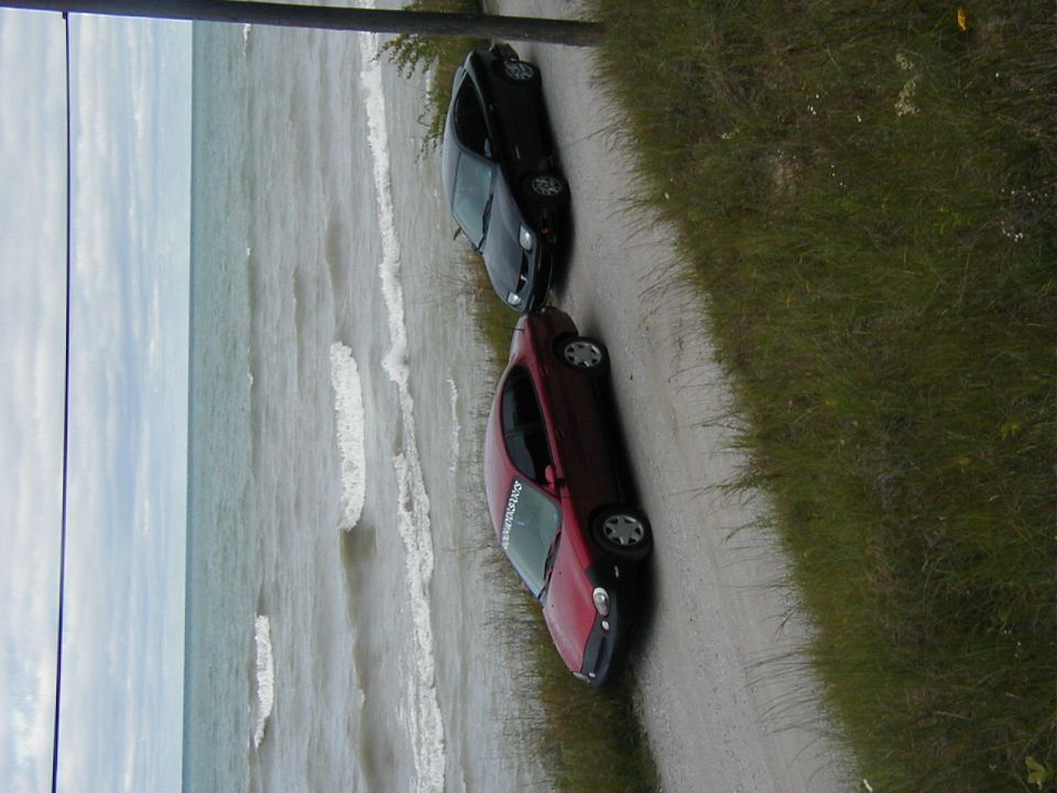 Two cars, a red one and a black one, parked on a dirt road next to the ocean