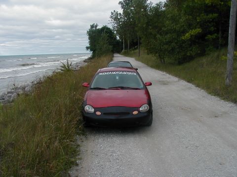1997 Ford Taurus - A red car with a black front bumper is parked on a gravel road next to a body of water.
