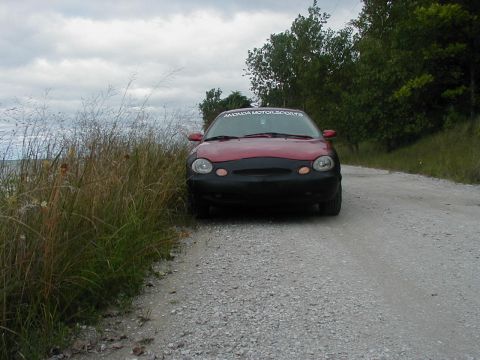 1997 Ford Taurus - A 1997 Ford Taurus is parked on a gravel road with tall grass to the left and trees in the background.