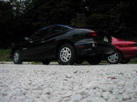 1997 Ford Taurus - Two cars are parked side-by-side on a gravel lot with a wooded area behind them.