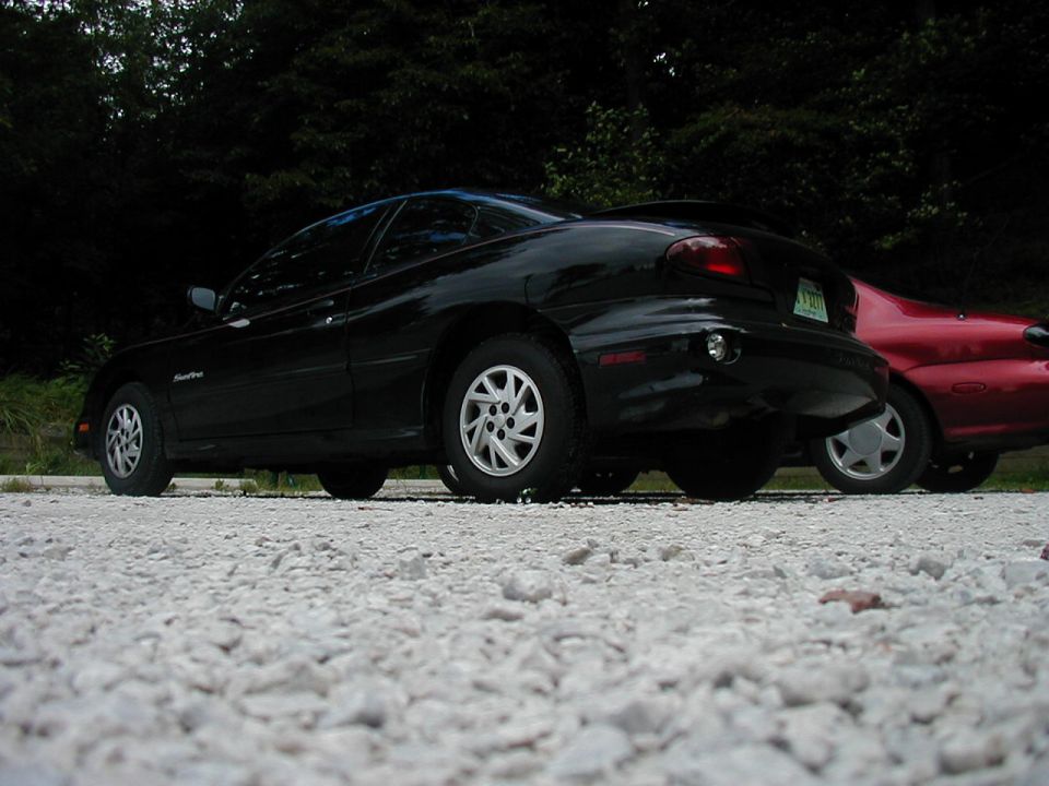 Black car parked next to a red car on a gravel surface with trees in the background