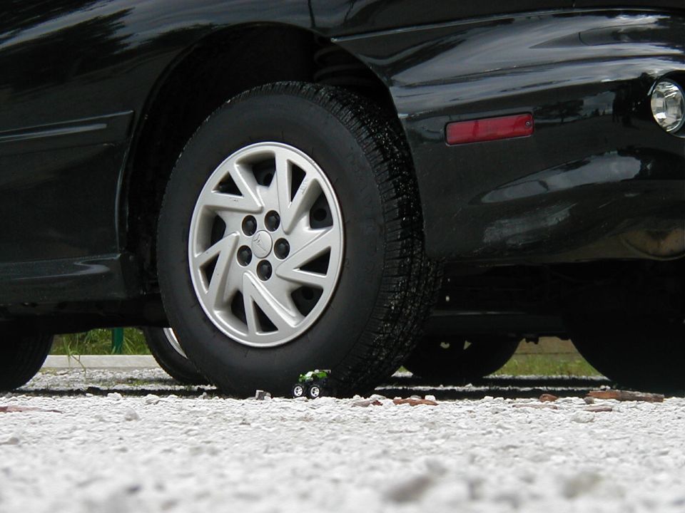 Tiny green toy car parked under the wheel of a black car
