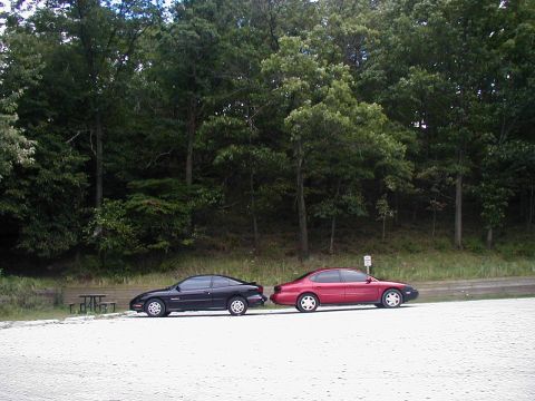 1997 Ford Taurus - Two cars, a red 1997 Ford Taurus and a black coupe, are parked side-by-side on a gravel lot with trees in the background.