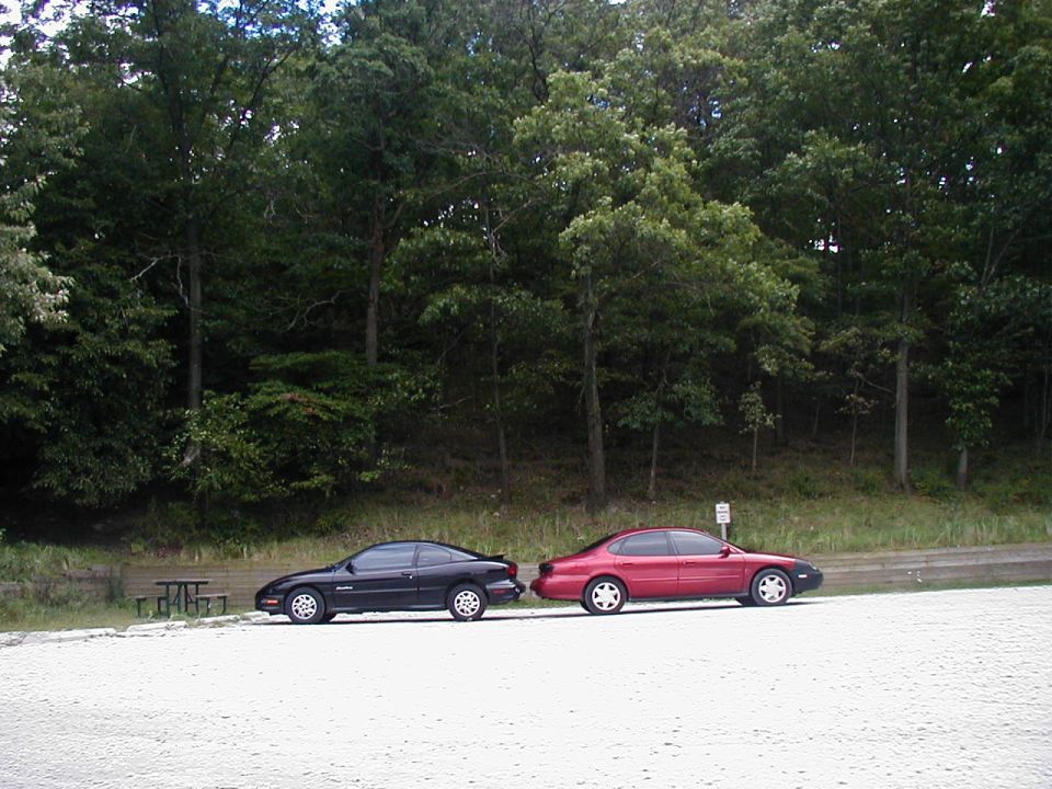Red 1997 Ford Taurus parked on a gravel lot next to a black coupe
