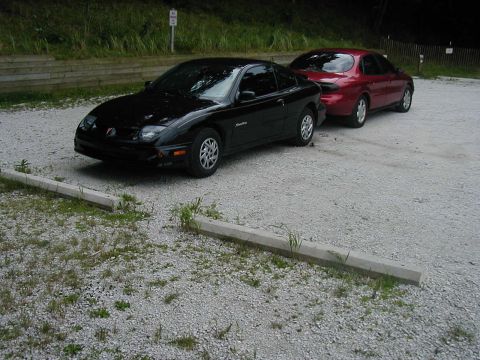 1997 Ford Taurus - Two cars, a black coupe and a red sedan, are parked on a gravel lot.