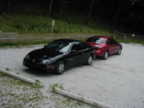1997 Ford Taurus - Two cars, a black coupe and a red sedan, are parked side-by-side in a gravel lot.