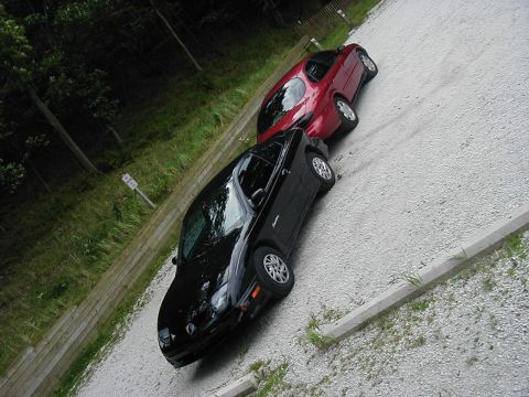 1997 Ford Taurus - Two cars are parked on a gravel surface with trees and a fence in the background.