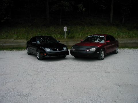 1997 Ford Taurus - Two sedans are parked on a gravel lot with trees in the background.
