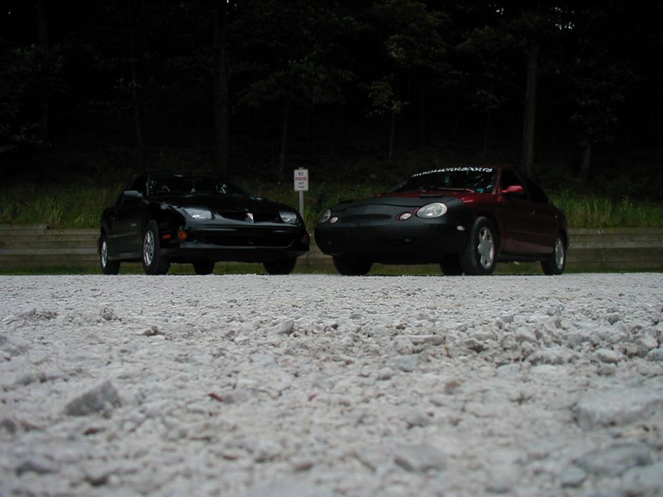 Two cars, a black Pontiac Firebird and a red car with a black cover, parked side-by-side on a gravel lot