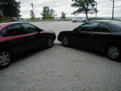 1997 Ford Taurus - A red sedan and a black coupe are parked nose-to-nose in a gravel lot, with a body of water visible in the background.