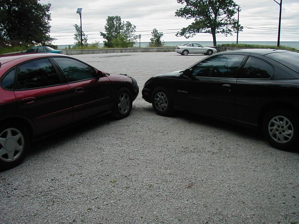 Two cars parked facing each other on a gravel lot near a body of water