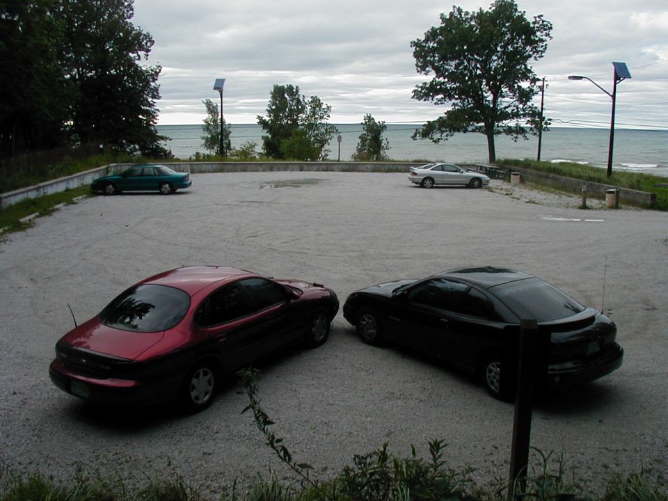 Maroon and black coupes parked in gravel lot near water; one green sedan in background