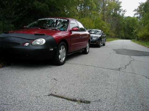1997 Ford Taurus - A red 1997 Ford Taurus sedan is parked on the side of a road, with another car behind it.