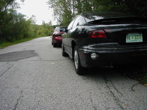 1997 Ford Taurus - Two cars are parked on the side of a road, one black coupe and a red car behind it.
