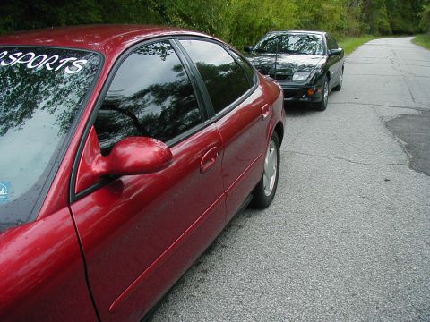 1997 Ford Taurus - Two cars are parked on a paved road surrounded by trees and greenery.