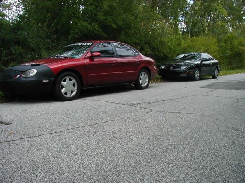 1997 Ford Taurus - Two sedans, a red Ford Taurus and a black car, are parked on the side of a paved road with trees in the background.