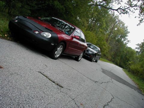 1997 Ford Taurus - A red 1997 Ford Taurus, partially covered by a car bra, is parked on the side of a road with another vehicle following behind.