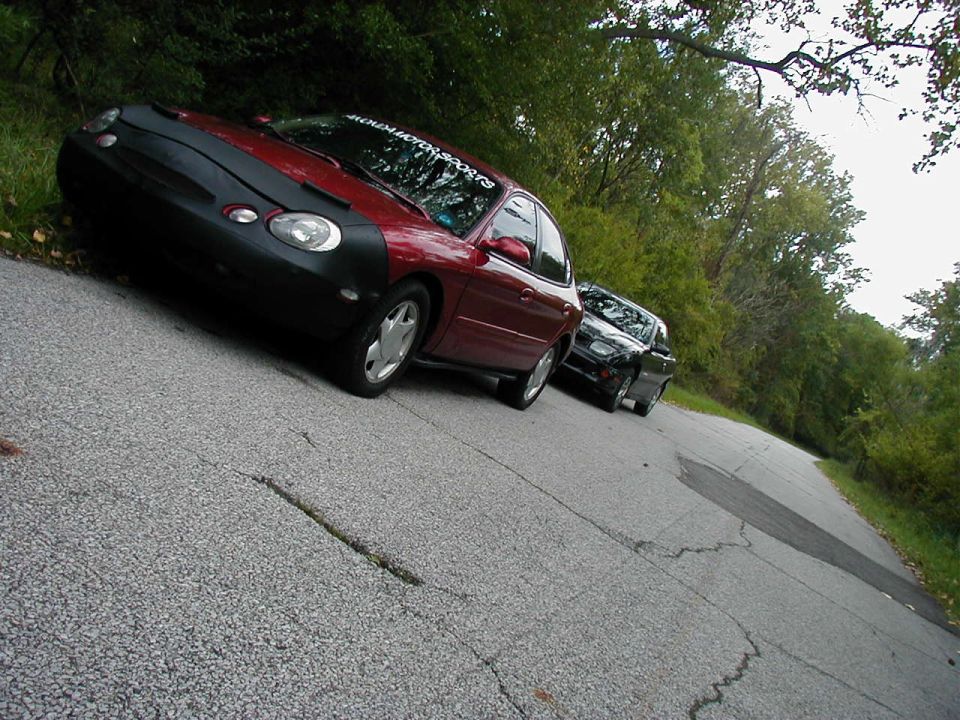 Red 1997 Ford Taurus with car bra, parked on a road with another car behind it
