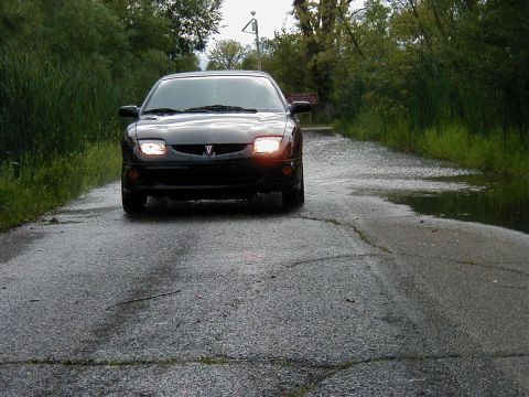 1997 Ford Taurus - A black Pontiac Sunfire is seen driving down a wet, cracked road surrounded by greenery.