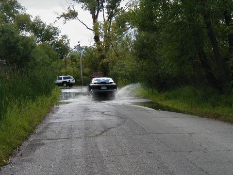 1997 Ford Taurus - A dark sedan drives through a flooded road, creating a large spray of water as a white SUV waits behind.