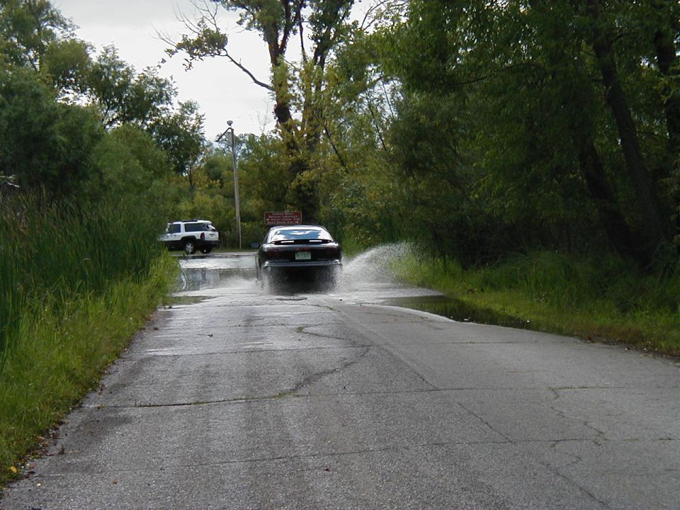 Dark sedan driving through water, kicking up spray. White SUV visible in background