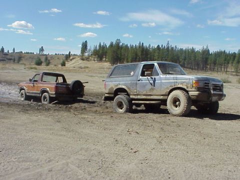1989 Ford Bronco - Two Ford Broncos navigate a muddy, off-road terrain under a partly cloudy sky.